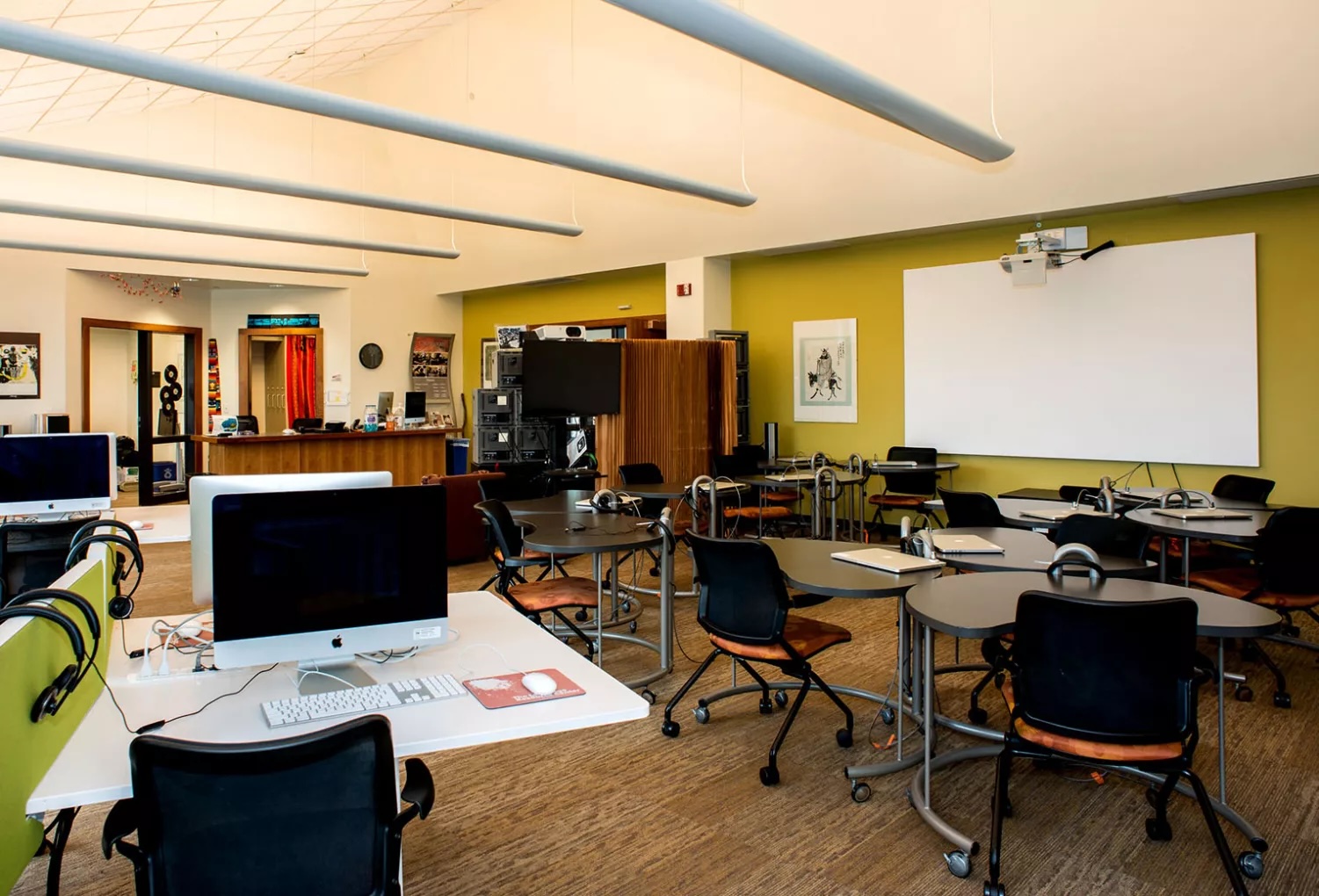 The Language Center main room with multiple study tables on wheels, computers on fixed tables, a white board screen on the wall below a projector. In the distance is the front desk.