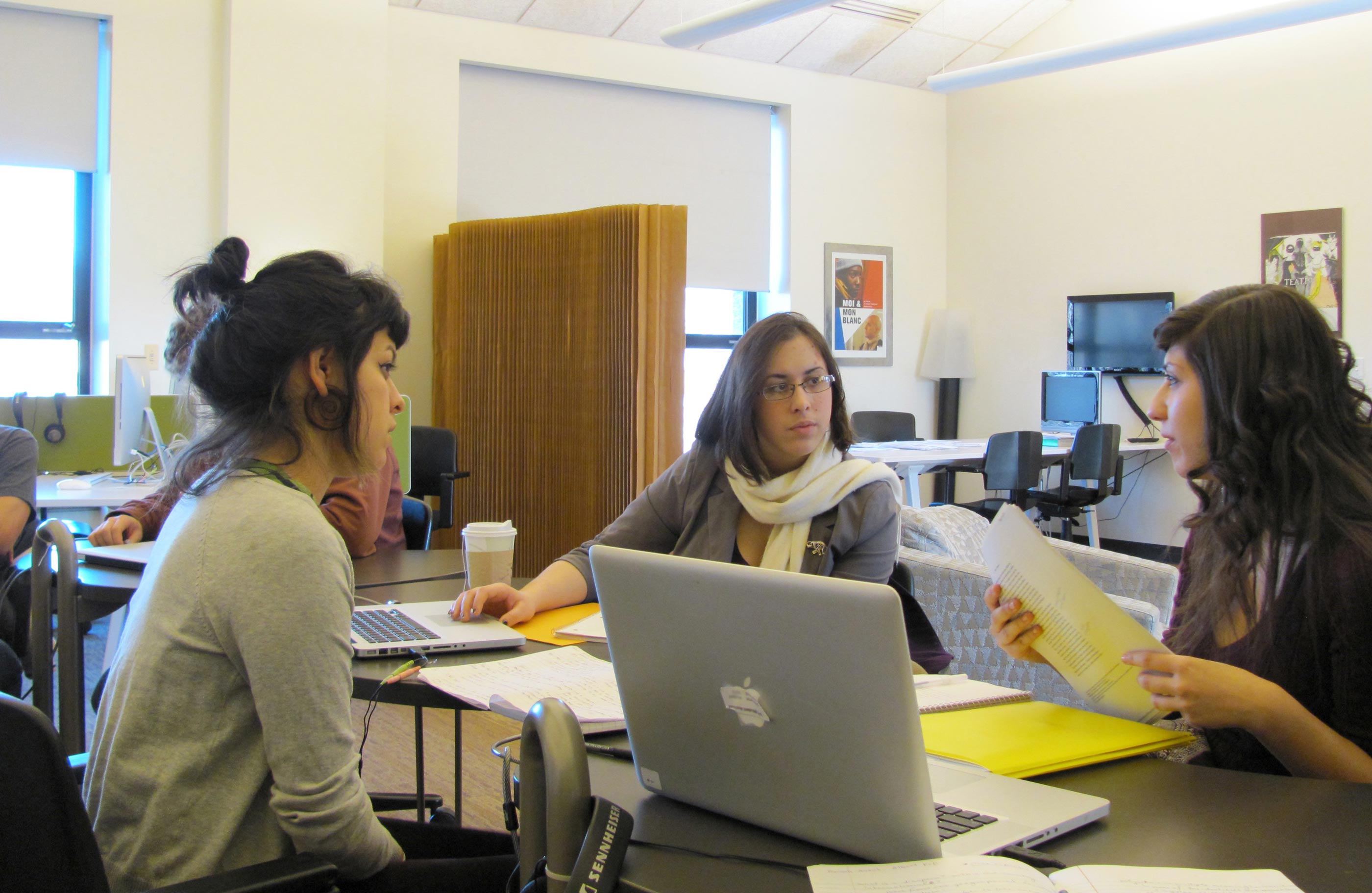 Three students sitting at desks, in a discussion. Laptop computers and notebooks sit on the desks. There are large widows in the back of the room and a collapsable noise and privacy wall in the background.